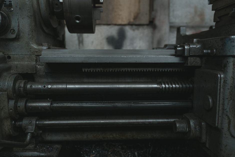 Close-up of a large industrial machine with metallic components, including a gear rack and linear guide rods, situated in a workshop or manufacturing environment. The machinery appears to have a dark,